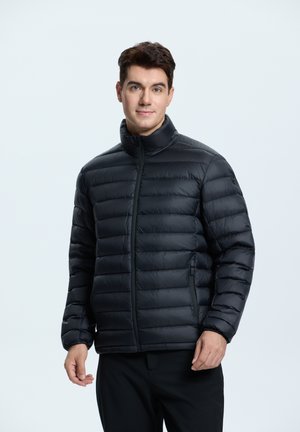 Zwarte pufferjack met een hoge kraag, gekleed ontwerp, voorkantsluiting met ritssluiting en zijzakken, gedragen over zwarte broeken. Gladde textuur.
