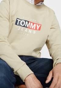 Felpa beige con scollo rotondo, caratterizzata da un grande logo "TOMMY JEANS" multicolore. Polsini e orlo a costine, indossata con jeans in denim blu scuro.