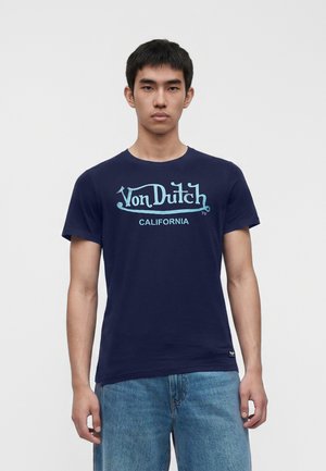 Giovane uomo che indossa una T-shirt blu navy Von Dutch California e jeans azzurri, in piedi davanti a uno sfondo bianco semplice.