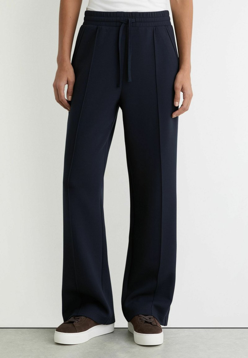 Henkilö, joka käyttää tummansinisiä, leveälahkeisia housuja etuvahvistuksilla, joustavalla vyötäröllä, jossa on kiristysnauha, ja ruskeita lenkkareita, joissa on valkoiset pohjat.