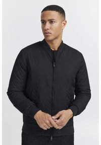 Giacca bomber nera realizzata in materiale liscio e imbottito con zip frontale, polsini e orlo a costine; presenta tasche laterali e un colletto rotondo.