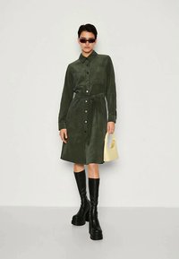 Γυναίκα φορώντας ένα σκούρο πράσινο φόρεμα με κουμπιά και ζώνη, μαύρες πλατφόρμες μέχρι το γόνατο, γυαλιά ηλίου και κρατώντας μια μικρή μπεζ τσάντα.