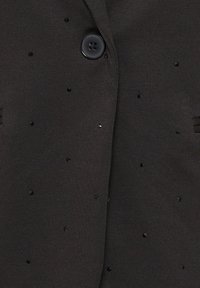 Must mustrikardigan, millel on tume sädelevad sekki, mis on hajutatud kangale, ja suur must nupp krae lähedal.