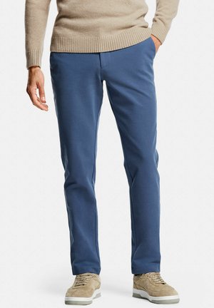 Homme portant un pull beige, un pantalon droit bleu et des baskets en daim beige, debout avec une main dans la poche sur un fond blanc.