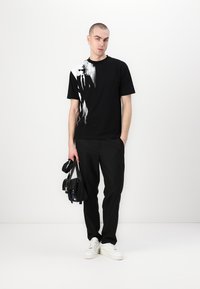 Črna majica s grafičnim tiskom na eni strani, v kombinaciji s črnimi hlačami in belimi supergami. Model drži črno torbo.