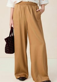 Brede broek in een lichtbruine stof met een elastische tailleband. Zakken zichtbaar aan beide zijden, gecombineerd met een kleine, donkerpaarse tas.