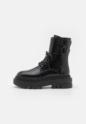 Black leather ankle boot with a round toe, lace-up front, and two buckle straps on the cuff. Chunky rubber sole with treaded design.