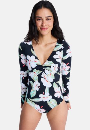 Žena v čiernych plavkách s dlhým rukávom s veľkým bielo-ružovým kvetinovým vzorom stojí a mierne sa usmieva do fotoaparátu.