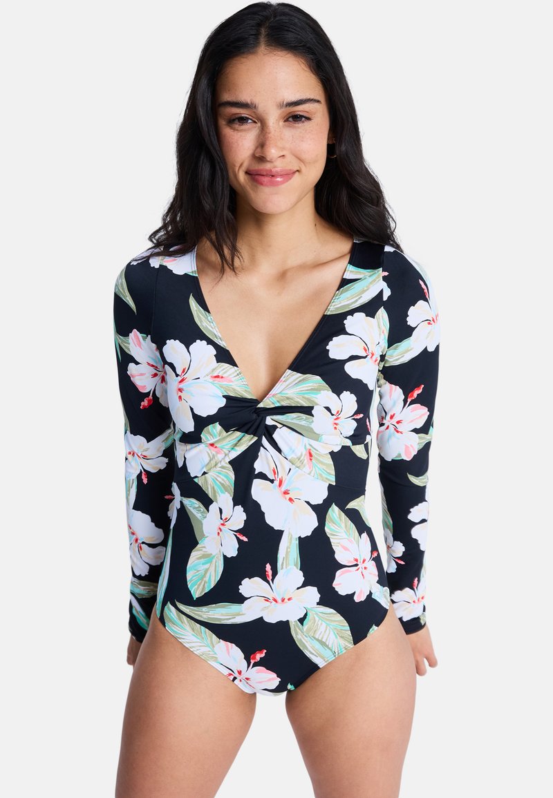 Γυναίκα που φοράει μαγιό μακρυμάνικο μαύρο με μεγάλο λευκό και ροζ λουλουδάτο σχέδιο, στέκεται και κοιτάζει προς την κάμερα, χαμογελώντας ελαφρώς.