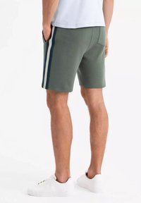 Shorts verts en matière douce avec des rayures verticales blanches et marines. Dotés de poches latérales et d'une seule poche arrière. Portés avec des baskets blanches.