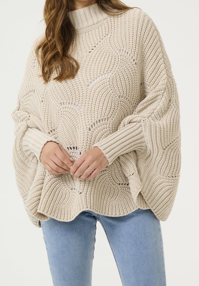 Beige strikket poncho med åbent vævemønster, høj hals, ribstrikkede ærmekanter og afslappet pasform, båret over lyseblå jeans.