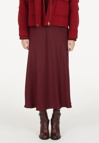 Frau in einem bordeauxfarbenen Midikleid, passenden Leder-Ankle-Boots und einer roten Zip-Jacke mit Fronttaschen.