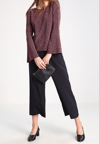 Top lungo a maniche lunghe bordeaux glitterato con maniche svasate, abbinato a pantaloni neri a gamba larga e scarpe nere senza lacci, mentre tiene in mano una borsa a tracolla nera.