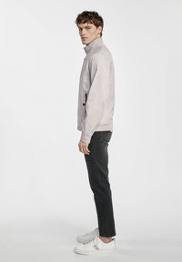 Jasnoszara kurtka bombka z wysokim kołnierzem, zamkami błyskawicznymi w kieszeniach i ściągaczami przy mankietach, w zestawie z ciemnymi dżinsami i białymi trampkami.