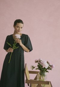 Vrouw in een zwart met witte stippen jurk, met een roze bloem in haar hand, staande naast een houten stoel met een vaas met roze pioenen, op een roze achtergrond.