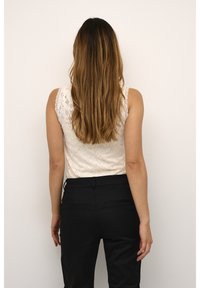 Haut sans manches en dentelle blanche avec des motifs élaborés, associé à un pantalon noir ajusté doté de poches arrière et d'une texture lisse.