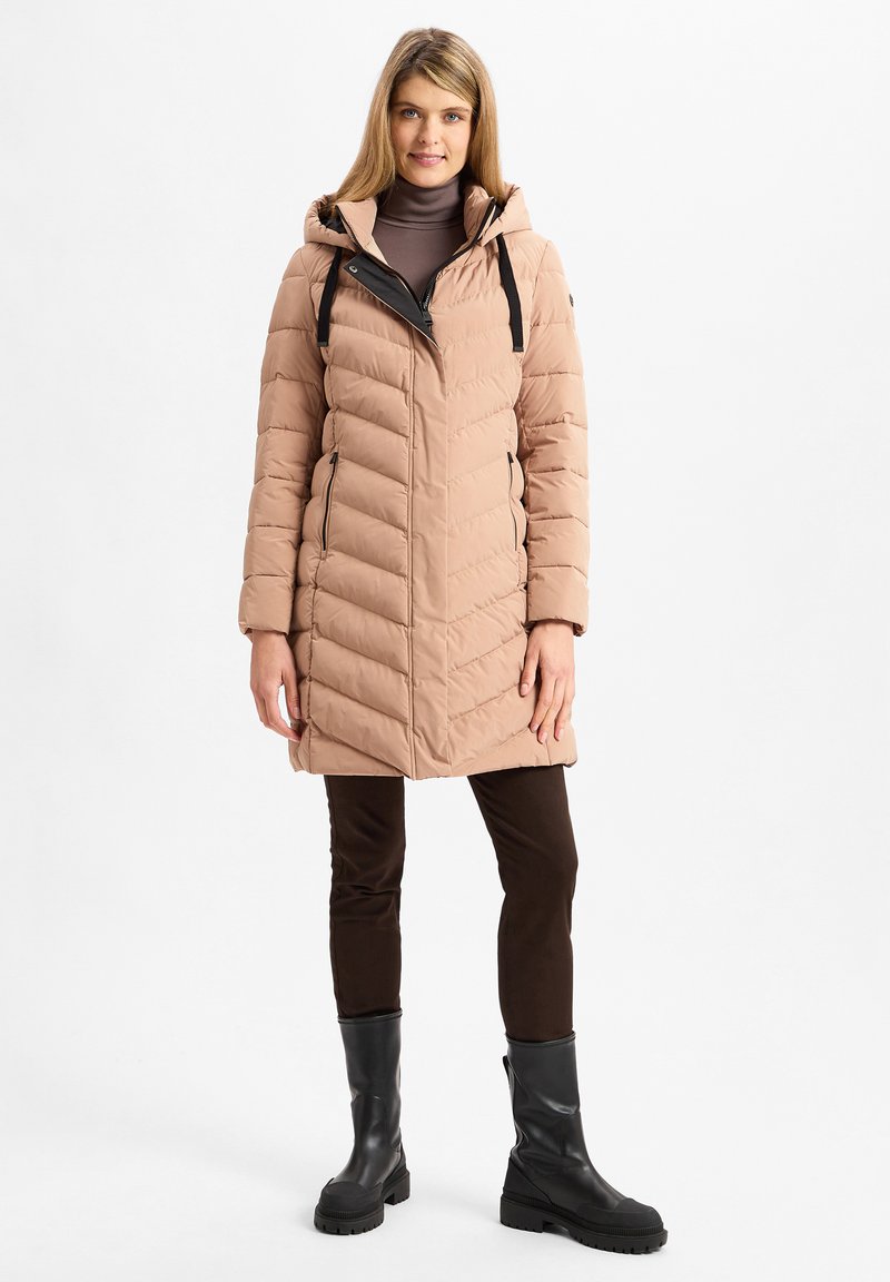 Puffermantel in hellem Beige mit Kapuze, ausgestattet mit einem Reißverschluss vorne, schwarzen Akzenten und einer gesteppten Textur. Dazu kombiniert mit braunen Hosen und schwarzen Stiefeln.