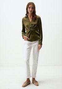 Blouse en satin de couleur vert olive foncé avec un décolleté en V, détail torsadé à la taille et manches longues ; assortie à un pantalon droit blanc et des chaussures beiges.