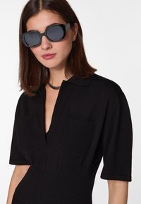 Robe tricot noir avec un décolleté en V, des manches courtes et deux poches poitrine. Les accessoires incluent de grandes lunettes de soleil noires et un collier choker sombre.