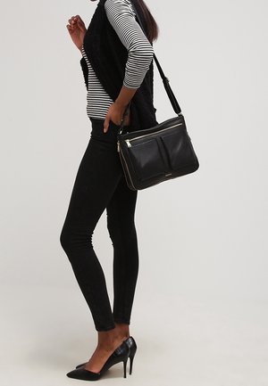 Sac à main en cuir noir avec deux poches zippées, accents de quincaillerie dorée. Haut à manches longues rayé, jean skinny noir et talons hauts.