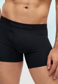 Мъжки торс с черни боксерки Abercrombie & Fitch, показващ талия, пъп с пиърсинг и дясна ръка, почиваща до тялото.