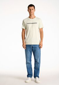 Camiseta de algodón de color verde claro con el texto "COPENHAGUE" en negro, combinada con jeans de mezclilla azules y zapatillas blancas. Diseño simple y casual.