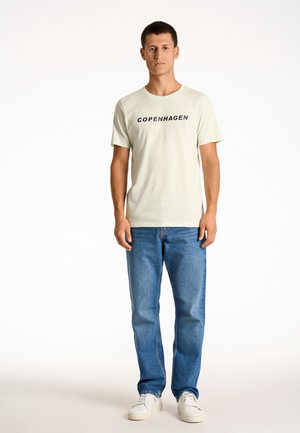 Camiseta de algodón de color verde claro con el texto "COPENHAGUE" en negro, combinada con jeans de mezclilla azules y zapatillas blancas. Diseño simple y casual.