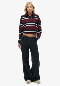 Kvinde står iført en cropped, stribet langærmet polo t-shirt, mørke vide bukser og lyse sneakers på hvid baggrund.