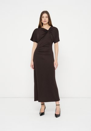 Femme portant une robe marron foncé à manches courtes et longueur cheville avec un détail torsadé au niveau de la poitrine, associée à des talons noirs à bride cheville, debout contre un mur blanc.