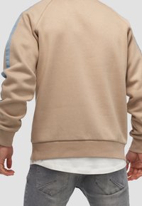 Beige sweatshirt med blå axelapplikationer, ribbad mudd och nederkant, samt mjukt material. Bärs över en vit skjorta och i kombination med grå jeans.