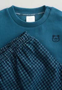 Turkusowy sweatshirt z emblematem misia i prążkowanym dekoltem w parze z ciemnoniebieskimi szortami w kratę z elastycznym pasem.
