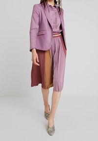 Donna che cammina in un blazer lilla, una blusa con volant, una gonna plissettata multicolore e tacchi a punta in pelle di serpente, su uno sfondo semplice.