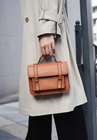 Brązowa skórzana torebka z górnym uchwytem, z klapką i detalami w postaci przeszyć. Trzymana w dłoni, w połączeniu z beżowym płaszczem trench.