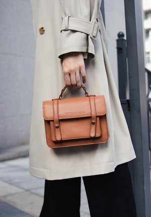 Bruine leren handtas met een bovenhandvat, klepsluiting en stiksels. Veroordeeld in een hand, gecombineerd met een beige trenchcoat.