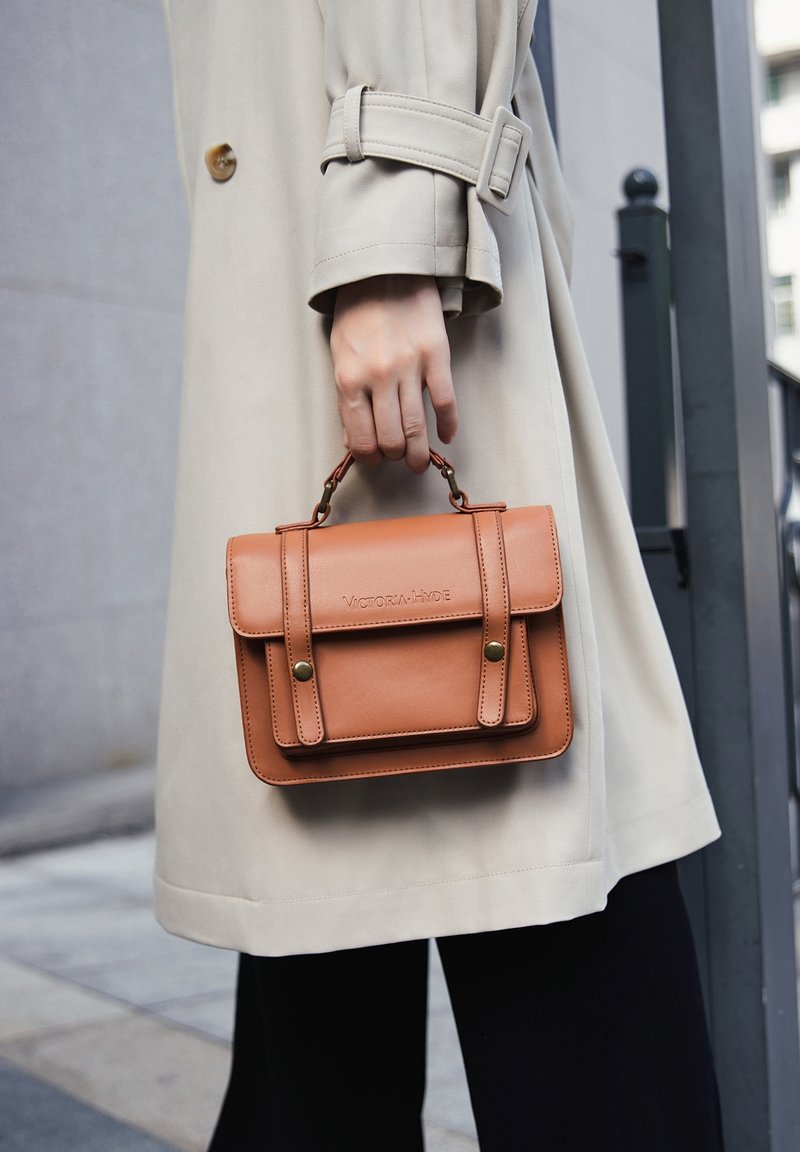 Brązowa skórzana torebka z górnym uchwytem, z klapką i detalami w postaci przeszyć. Trzymana w dłoni, w połączeniu z beżowym płaszczem trench.
