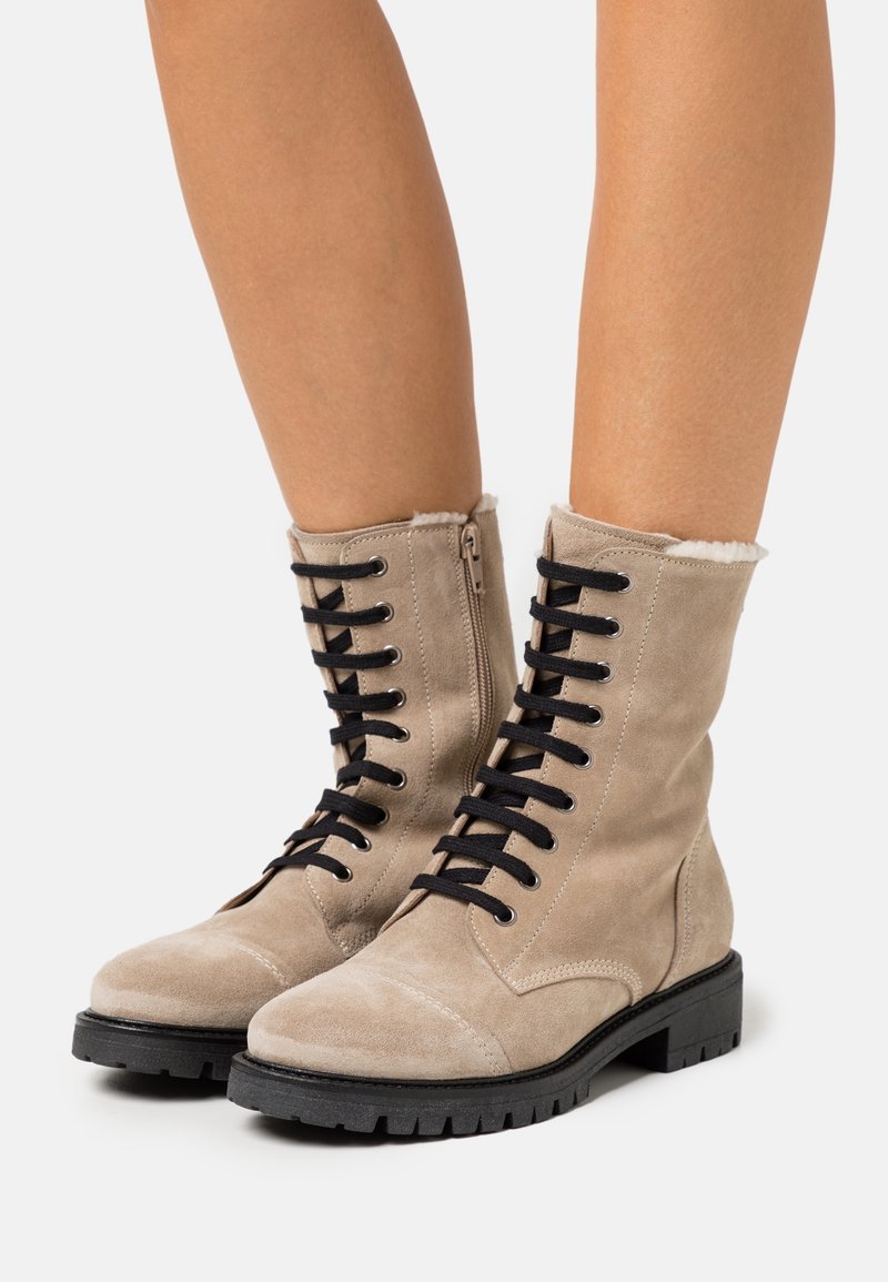 Béžové semišové kotníkové boty s černými tkaničkami, bočním zipem a kožešinovou podšívkou na manžetě; robustní černá gumová podrážka, vojenský styl.