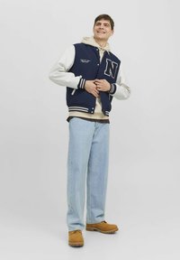 Marine varsity jack met witte mouwen, gestreepte boorden en "N" logo; beige hoodie eronder; lichte denimbroek; tan schoenen.