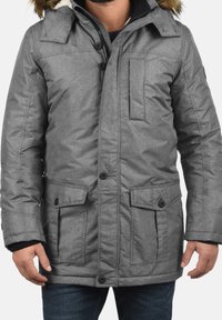 Homme portant une doudoune grise avec capuche bordée de fourrure, fermeture par boutons et zip, et poches à rabat, debout avec les bras détendus.