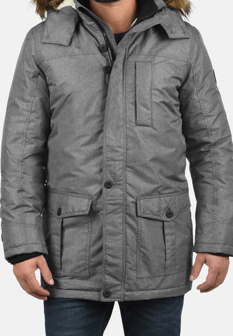 Homme portant une doudoune grise avec capuche bordée de fourrure, fermeture par boutons et zip, et poches à rabat, debout avec les bras détendus.