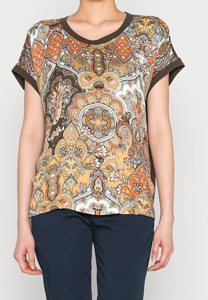 Femme portant un chemisier à manches courtes avec un galon marron et un motif paisley complexe orange, beige et blanc, associé à un pantalon foncé.