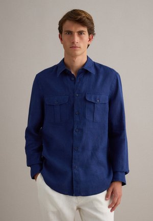 Joven con camisa de manga larga azul marino abotonada con bolsillos en el pecho y pantalones blancos, posando frente a un fondo liso beige.