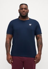 Tricou din bumbac moale, cu mânecă scurtă, de culoare bleumarin, cu guler rotund și detaliu logo alb pe piept; design simplu, fără modele.