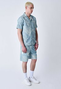 Camisa de manga corta azul claro y pantalones cortos a juego que presentan patrones de hojas blancas. El conjunto incluye zapatillas y calcetines blancos.