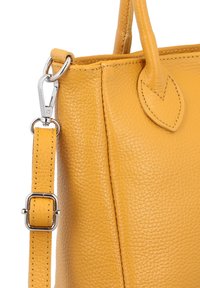 Kollase nahast käekott, millel on tekstureeritud pind, eemaldatava rihma ja hõbedaste detailide aktsentidega. Küljel on lehekujuline detail.