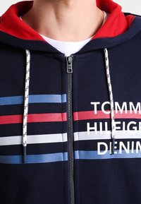 Marinblå zip-up hoodie med röd huva, med vita, röda och blå ränder. Tryckt text "TOMMY HILFIGER DENIM" i vitt. Texturerat tyg.