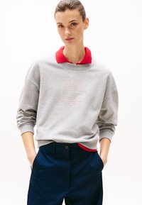 Szara bluza z czerwonym kołnierzykiem, z nadrukowanym logo. Design jest casualowy, z luźnym krojem i ściągaczami na rękawach, noszona z granatowymi spodniami.