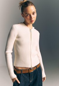 Biały kardigan z zapięciem na zamek i kapturem, o dopasowanym kroju, z długim rękawem i subtelną fakturą. W połączeniu z ciemnymi jeansami i brązowym paskiem.