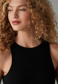Top nero senza maniche con collo alto. Tessuto liscio e elasticizzato con un design aderente. Capelli ricci e sciolti che incorniciano il viso.