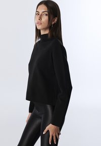 Mladá žena v černém dlouhém topu s dlouhým rukávem a lesklých černých leginách, stojící na boku proti jednobarevnému světlému pozadí.