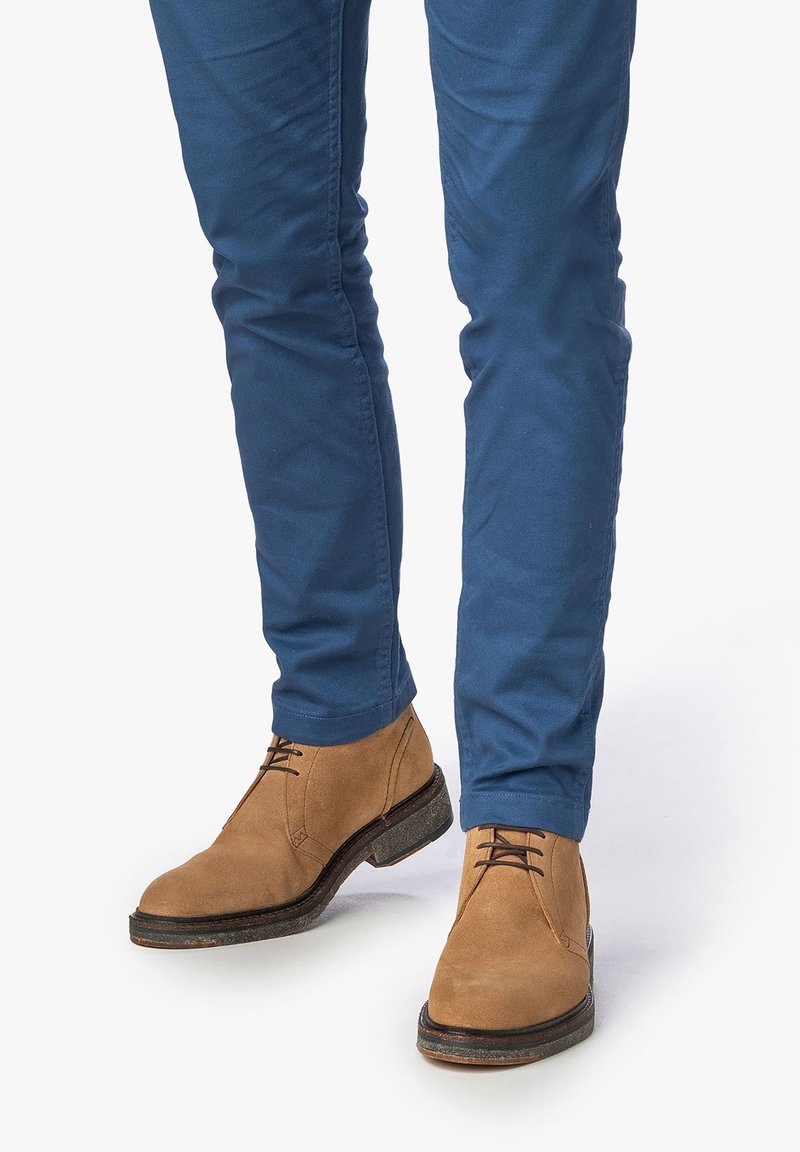 Botas de ante marrón hasta el tobillo con suelas de goma negras; combinadas con pantalones ajustados azules. Las botas tienen punta redondeada y un cordón mínimo.